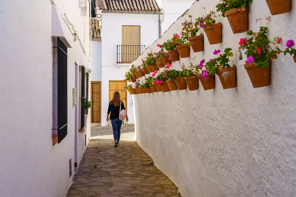 Mujer paseando por un callejón estrecho lleno de macetas y flores en un pueblo blanco de Andalucía, ideal para rutas por el sur de España.